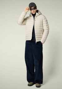 Uomo che indossa un giubbotto beige invernale, pantaloni navy a gamba larga, dolcevita nero, cappellino nero e occhiali da sole neri, mentre aggiusta gli occhiali da sole con una mano.