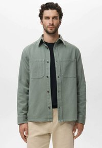 Grüne Baumwollhemdjacke mit Knopfverschluss, zwei Brusttaschen und gewickeltem Kragen, getragen über einem schwarzen Hemd.