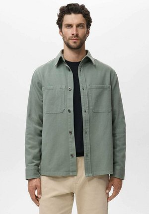 Green cotton shirt jacket featuring a button-down front, two chest pockets, and a collared neckline, worn over a black shirt.