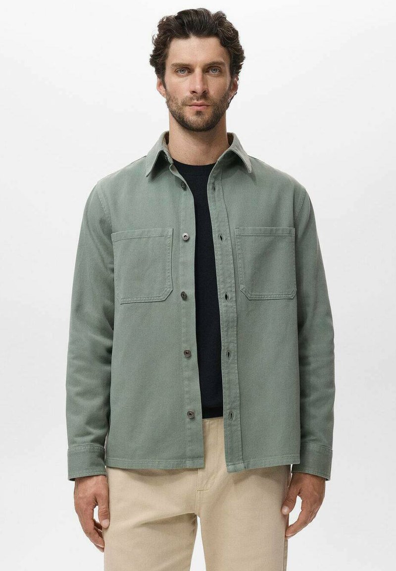 Grüne Baumwollhemdjacke mit Knopfverschluss, zwei Brusttaschen und gewickeltem Kragen, getragen über einem schwarzen Hemd.