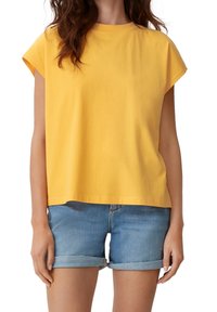 Kobieta ubrana w luźną, żółtą bluzkę z krótkim rękawem i jasnoniebieskie szorty dżinsowe z podwiniętymi nogawkami na białym tle.