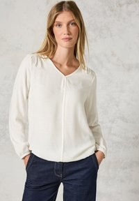 Vit långärmad blus med V-ringning, med strukturerade diamantmönster, knäppning med knappar och volangärmar. Kombinerad med mörka jeans.