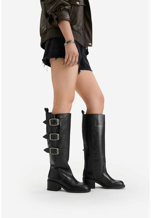 Nogi w czarnych skórzanych kozakach sięgających do kolan, z trzema klamrami na jednym bucie i gładkim wzorem na drugim, w połączeniu z czarnymi szortami i brązową kurtką.