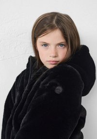 Chaqueta de piel sintética negra con capucha, que presenta una superficie texturizada y un parche con un logotipo redondo en el hombro izquierdo.
