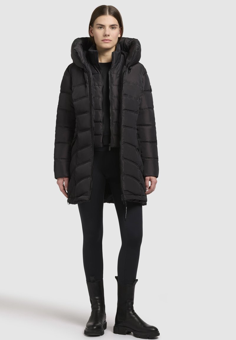 Femme portant un long manteau matelassé noir avec capuche, des leggings noirs et des bottes noires, se tenant devant un fond gris uni.