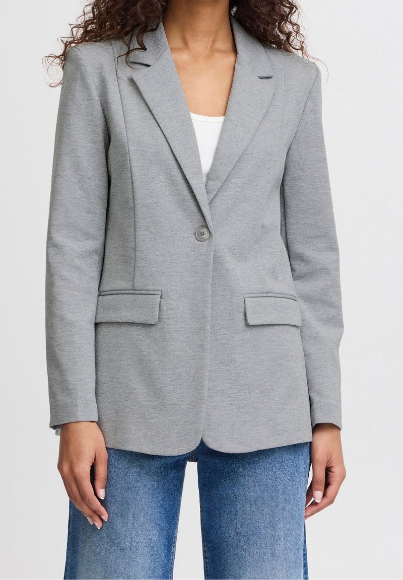 Femme portant un blazer gris clair à un bouton sur un haut blanc avec un jean bleu en denim, debout devant un fond uni.