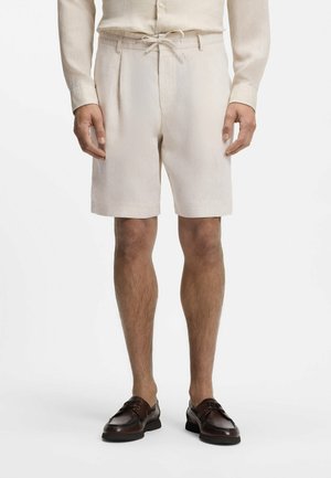 Mężczyzna w beżowych szortach z sznurkiem, beżowej koszuli z długim rękawem oraz ciemnobrązowych skórzanych mokasynach, stojący na białym tle.