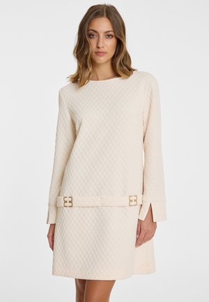 Vrouw draagt een crèmekleurige, doorgestikte jurk met lange mouwen en een bijpassende riem met gouden rechthoekige gespen.