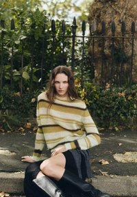 Maglione a righe giallo, bianco e marrone. Texture in maglia morbida con vestibilità comoda, abbinato a una gonna scura e stivali neri fino al ginocchio.