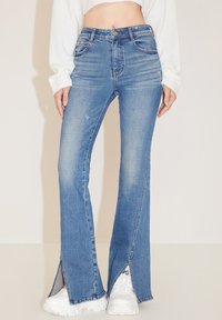 Jeans acampanados de cintura alta en denim azul claro, con aberturas laterales en el dobladillo y ligeras arrugas. Combinados con zapatillas blancas de suela gruesa.