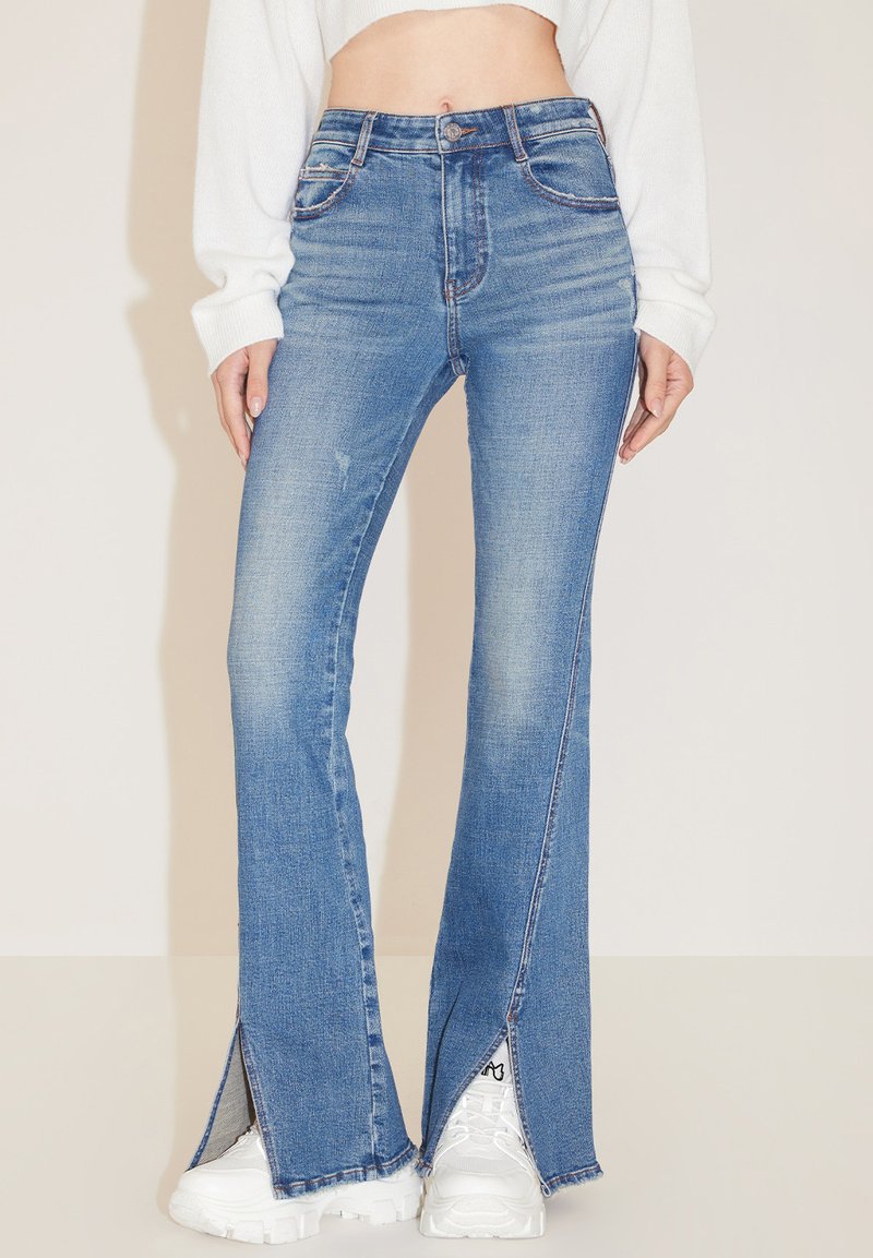Jeans acampanados de cintura alta en denim azul claro, con aberturas laterales en el dobladillo y ligeras arrugas. Combinados con zapatillas blancas de suela gruesa.