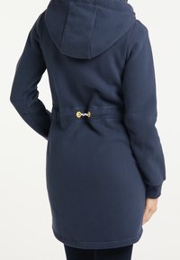 Marinblå luvtröja med dragsnodd i midjan, med en gul toggle. Mjuk material, långa ärmar och en avslappnad passform.