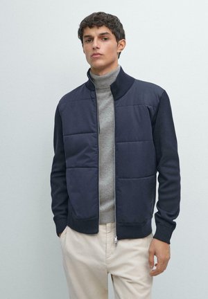 Giacca imbottita blu navy con chiusura a zip, polsini a costine e collo alto, indossata sopra un dolcevita grigio chiaro. Dettagli in tessuto strutturato.