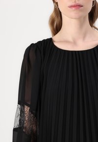 Abito nero plissettato con accenti di pizzo trasparente sulle maniche. Collo rotondo e tessuto morbido e fluido creano una silhouette elegante.