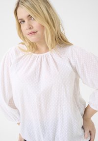 Blouse blanche à manches longues avec un col froncé, tissu texturé avec de petits pois, et poignets élastiques.