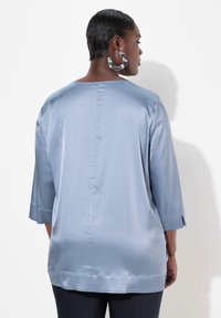 Top in satin azzurro chiaro con maniche a tre quarti, leggermente svasato e con spacchi laterali all'orlo. Texture liscia e design minimalista.