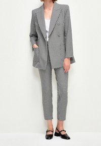 Costume à carreaux noir et blanc comprenant un blazer long avec des poches et un pantalon ajusté. Associé à des chaussures noires avec un détail de sangle.