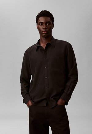 Junger Mann mit dunkler Haut und kurzen lockigen Haaren, trägt ein schwarzes, langärmliges Hemd mit Knöpfen, Hände in den Taschen, neutraler grauer Hintergrund.