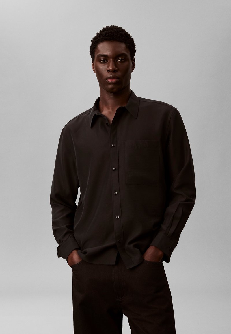 Jeune homme à la peau foncée et aux cheveux courts bouclés, portant une chemise noire à manches longues boutonnée, mains dans les poches, fond gris neutre.