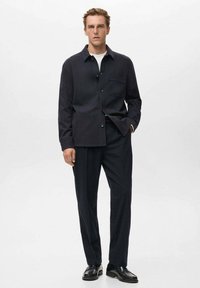 Giacca elegante di un blu scuro, con tasca sul petto, chiusura a bottone e pantaloni coordinati. Indossata sopra una maglietta bianca a collo tondo.