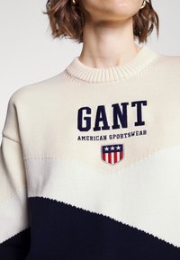 Pull en maille présentant un design à blocs de couleurs crème, marine et blanc avec un col côtelé. Orné du logo "GANT" et d'un blason américain.