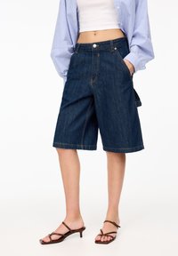 Bermudas en jean bleu foncé avec une coupe ample, dotées d'un bouton devant, de poches latérales et de surpiqûres contrastées.