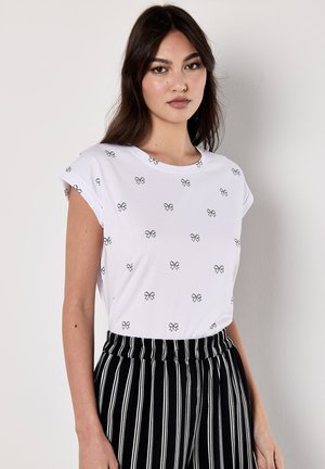 Modella che indossa una camicia bianca con motivi a farfalla nera e pantaloni a strisce nere, in posa su uno sfondo neutro.