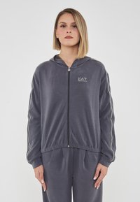 Felpa grigia con zip realizzata in tessuto liscio, con strisce laterali e cappuccio. Logo visibile sul petto con dettagli dell'elastico in vita.