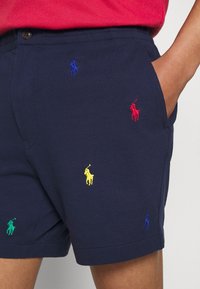 Bermudas azul marino fabricadas con un tejido texturizado, con logotipos de polo bordados en varios colores: rojo, azul, amarillo y verde. Bolsillo frontal visible.
