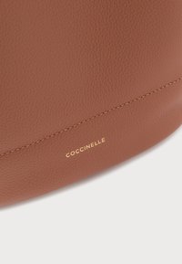 Καφέ δερμάτινη τσάντα χειρός με υφή στην επιφάνεια, που διαθέτει χρυσό λογότυπο "Coccinelle", διακριτικό ράψιμο και στρογγυλεμένο σχήμα.