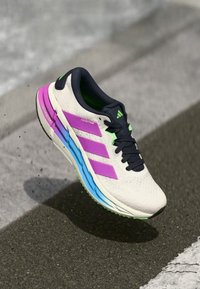 Λευκό αθλητικό παπούτσι Adidas με ροζ ρίγες και μπλε σόλα, αιωρούμενο πάνω από την άσφαλτο.