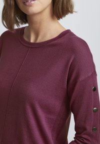 Stickad tröja i djup burgundy med rundad hals, med guldknappade detaljer på ärmarna och en slät textur.