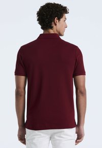 Bordeaux poloshirt met korte mouwen, een geribde kraag en zijsplitten. Gemaakt van een gladde, textuurrijke stof. Achteraanzicht weergegeven.