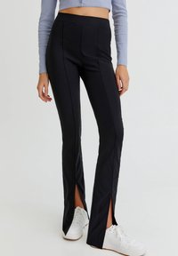 Pantalones ajustados negros con dobladillos partidos, confeccionados en una tela suave. Presentan una cintura alta y costuras verticales, combinados con zapatillas blancas.