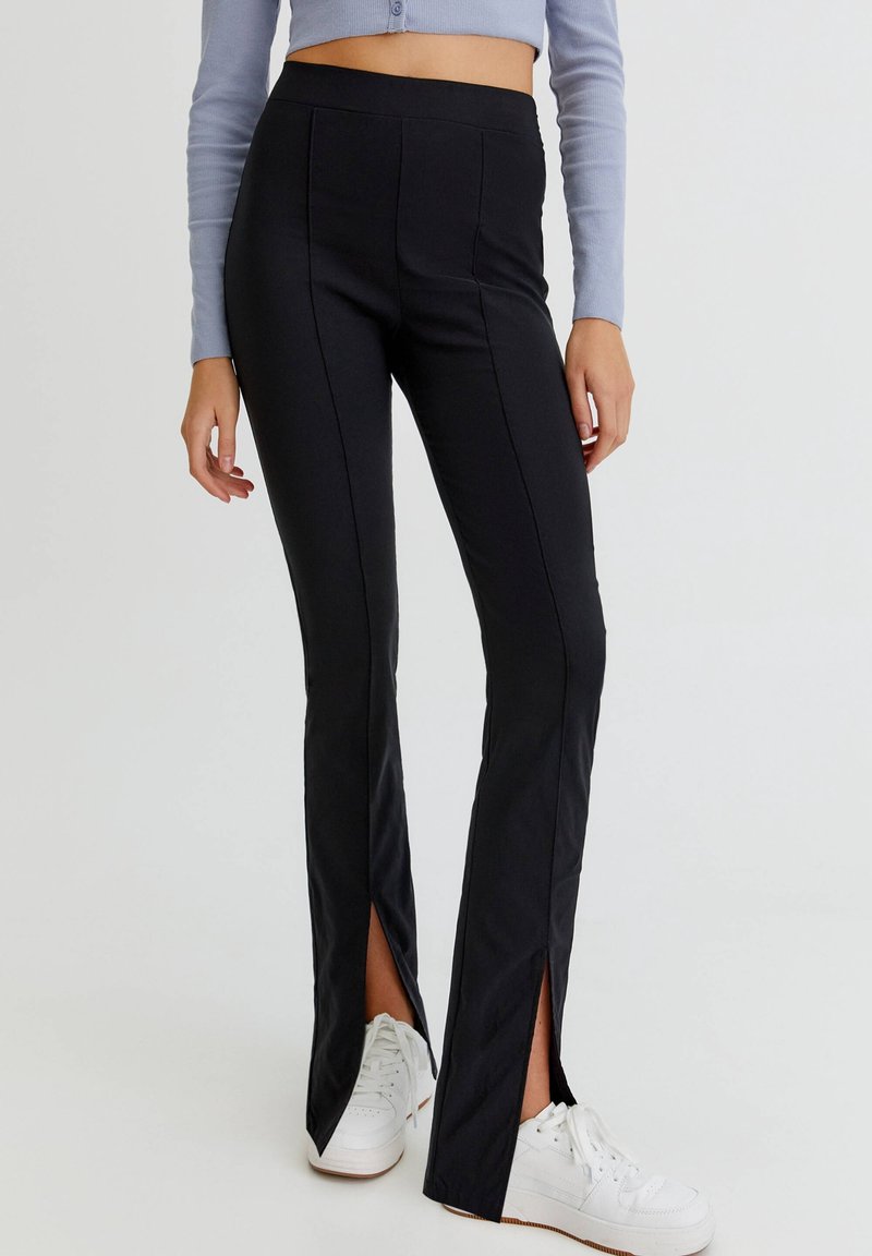 Pantalones ajustados negros con dobladillos partidos, confeccionados en una tela suave. Presentan una cintura alta y costuras verticales, combinados con zapatillas blancas.