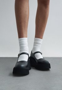 Πόδια που φορούν λευκές κάλτσες πλήρωσης σε συνδυασμό με μαύρα παπούτσια Mary Jane με πλατφόρμα, στέκονται σε γκρι πάτωμα με απλό φόντο.