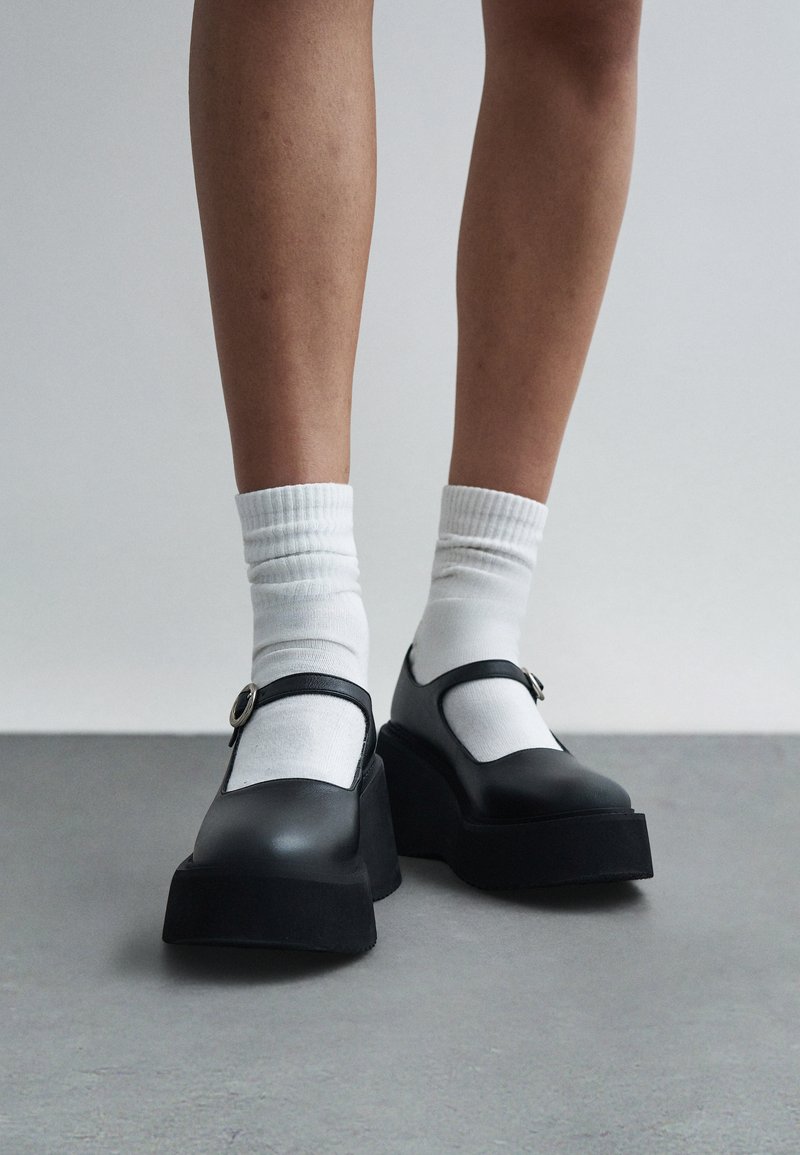 Πόδια που φορούν λευκές κάλτσες πλήρωσης σε συνδυασμό με μαύρα παπούτσια Mary Jane με πλατφόρμα, στέκονται σε γκρι πάτωμα με απλό φόντο.
