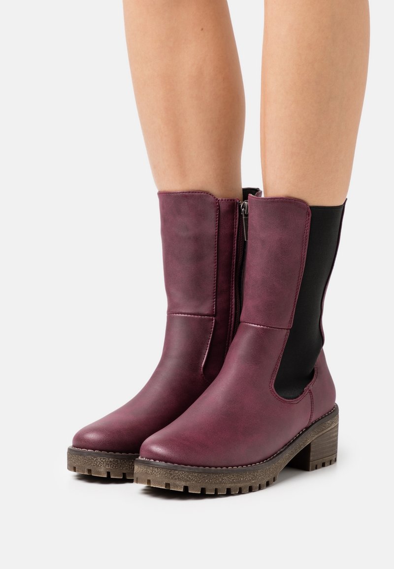 Bottes en cuir bordeaux mi-mollet avec talons épais et semelles marron texturées portées sur jambes nues, avec des panneaux latéraux élastiques noirs et des fermetures éclair latérales.