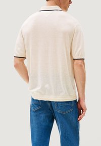 Polo beige con colletto, caratterizzata da una rifinitura scura al collo e alle maniche. Indossata con jeans blu, che mostrano una vestibilità comoda e una texture morbida.