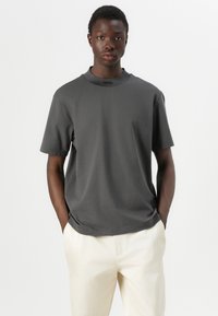 T-shirt gris à manches courtes en tissu lisse avec un col rond. Présente un logo de marque discret sur le col. Porté avec un pantalon de couleur claire.