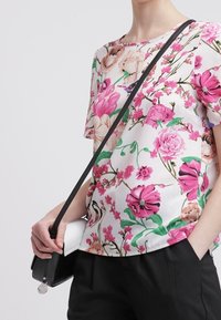 Kwiatowa bluzka z krótkim rękawem w różowe i zielone wzory na białym tle, zestawiona z czarną torebką na ramię i czarnymi spodniami.