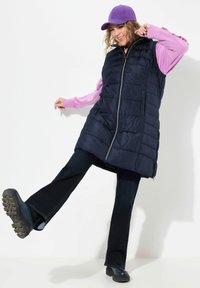 Marinblå puffer väst med dragkedja framtill, bärs över en ljuslila långärmad skjorta. Mörka utsvängda jeans och svarta klumpiga skor. Lila keps.
