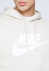 Béžová mikina s kapucí s bílým logem Nike a švem. Má kapuci se stahovací šňůrkou a texturovanou látku. Pohled zepředu, oříznuto u ramen.