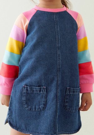 Kind trägt ein blaues Jeanskleid mit Vordertaschen über einem langärmeligen Hemd mit rosa, pfirsichfarbenen, violetten, gelben, blauen und roten Streifen.