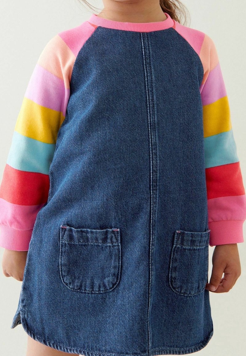 Enfant portant une robe en denim bleu avec des poches avant, par-dessus un t-shirt à manches longues rayé de rose, pêche, violet, jaune, bleu et rouge.