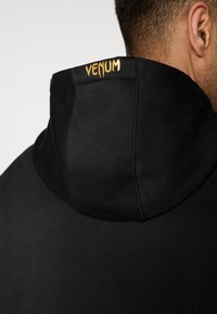 Μαύρο φούτερ με κουκούλα και ψηλό γιακά με υφή υφάσματος. Διαθέτει μικρό χρυσό λογότυπο "Venum" στο πίσω μέρος του λαιμού.