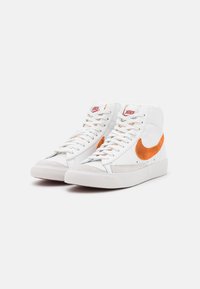 Vita högskor med texturerat läder, orange mocka swoosh och en gummisula. Snörningen är vit med röd branding på tungan.