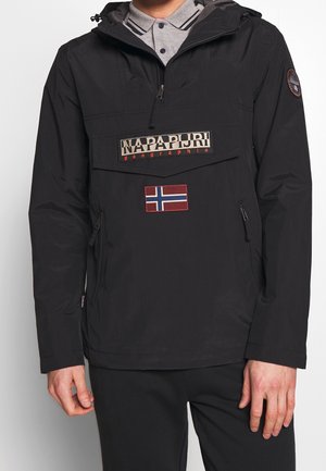 Personne portant une veste Napapijri noire avec une fermeture éclair à mi-hauteur, une poche avant à rabat et un écusson du drapeau de la Norvège sur la poitrine.