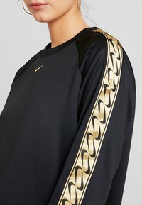 Svart sweatshirt med guld Nike-logga och guldaccentuerade mönstrade ränder på ärmarna; axlarna har en slät textur och ribbad halsringning.