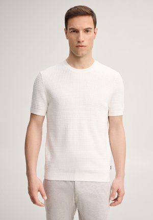 Uomo in piedi rivolto in avanti, che indossa un maglione bianco a maniche corte con trama e pantaloni grigio chiaro, con sfondo neutro.